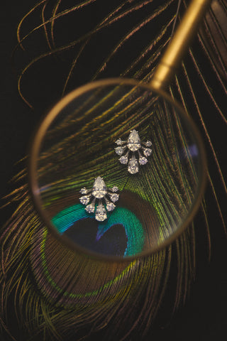THE HERA FRONTBACK EARRINGS ON A PEACOCK FEATHER, WITH A MAGNIFYING GLASS OVER THEM