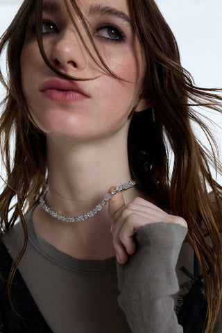 A MODEL LOOKING UPWARD WITH HER HAIR BLOWING IN FRONT OF HER FACE, AS SHE WEARS A GRAY LONG SLEEVE AND THE SUPERNOVA COLLAR NECKLACE, WITH HER THUMB LACED UNDER THE JEWELRY