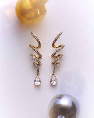 NADRI’S MUSE TWIRL LINEAR DROP EARRINGS LAID OUT AGAINST A WHITE BACKGROUND WITH PLAYFUL TOPS SPINNING AROUND IT
