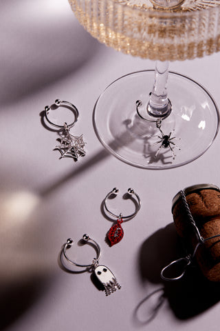 Set of four Halloween-themed wine charms on a table next to a wine glass. Charms include a ghost, spider, spiderweb, and red lips with vampire fangs.
