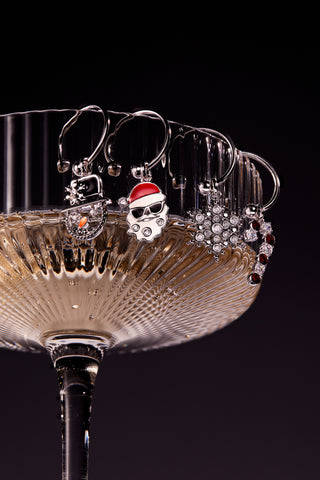 Wine glass with Christmas-themed wine charms on a black background.  The charms include a santa clause wearing sunglasses, a snowflake, a candy cane, and a snowman