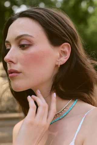 SIDE PROFILE OF A MODEL OUTSIDE WEARING THE SMALL PAVE HUGGIE HOOP EARRINGS AND LAYERED NECKLACES, WITH HER HAND LIFTED UP TO HER CHIN