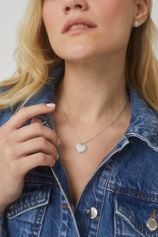 CLOSE UP IMAGE OF MODEL WEARING OUR GIFTED PAVE CZ HEART PENDANT IN RHODIUM PLATING WITH A DENIM JACKET.  SHE IS ALSO WEARING MATCHING EARRINGS AND HER HAND IS VISIBLE AND HOLDING THE COLLAR OF HER JACKET.