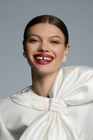 LONDON LARGE CLUSTER PEARL EARRINGS ON MODEL. SHE IS SMILING BIG WITH RED LIPSTICK ON AND HER HAIR BACK IN A SLICK BUN. SHE IS LOOKING DIRECTLY INTO THE CAMERA WITH BOTH EARRINGS VISIBLE. WEARING A WHITE TOP WITH A BOW IN A BRIDAL STYLE. 