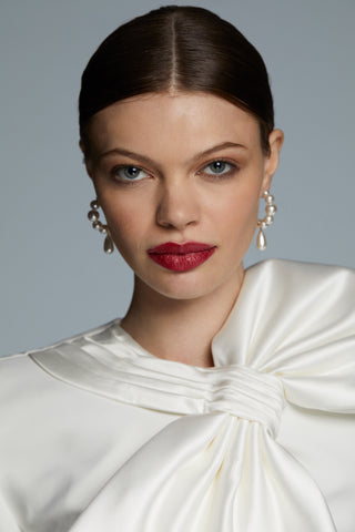 MODEL WEARING LONDON PEARL DROP C HOOP EARRINGS FACING DIRECTLY INTO THE CAMERA WITH BOTH EARRINGS VISIBLE WITH HER HAIR IN A SLICK BACK BUN AND RED LIPSTICK. 