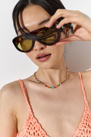 CLOSE UP OF A MODEL WEARING SUNGLASSES, THE FRIEDA FULL COLLAR NECKLACE, AND AN ORANGE TOP, WITH HER HAND RAISED UP TO HER GLASSES