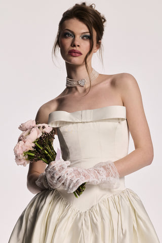A MODEL LOOKING AWAY FROM THE CAMERA, HOLDING A BOUQUET OF FLOWERS WHILE WEARING THE MATERIAL PEARL CHOKER NECKLACE, A WHITE DRESS, AND WHITE LACE GLOVES.