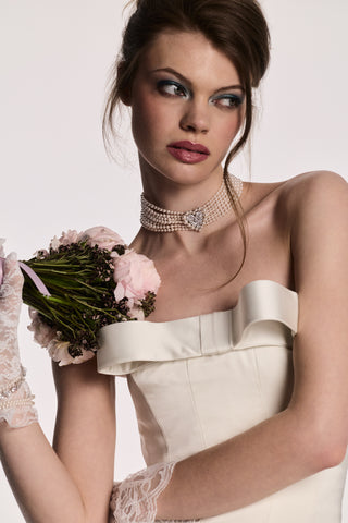 A MODEL LOOKING AWAY FROM THE CAMERA, HOLDING UP A BOUQUET OF FLOWERS TO HER SHOULDER WHILE WEARING THE MATERIAL PEARL CHOKER NECKLACE AND WHITE LACE GLOVES.