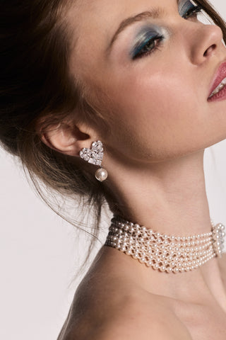 SIDE PROFILE OF A MODEL LOOKING UPWARD WEARING MATERIAL PEARL HEART DROP EARRINGS AND THE MATERIAL PEARL CHOKER NECKLACE, PAIRED WITH AN UPDO AND BLUE EYESHADOW.