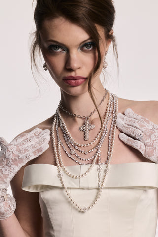 A MODEL LOOKING AT THE CAMERA WEARING THE MATERIAL PEARL DRAMA SWAG NECKLACE AND THE MATERIAL PEARL CROSS PENDANT NECKLACE, WITH HER HANDS UP TO HER CHEST FRAMING THE STACK.