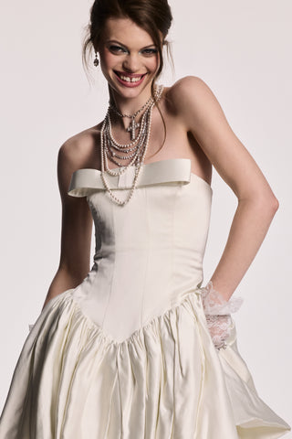 A MODEL SMILING AT THE CAMERA WEARING THE MATERIAL PEARL DRAMA SWAG NECKLACE AND THE MATERIAL PEARL CROSS PENDANT NECKLACE, STYLED WITH A WHITE DRESS AND LACE GLOVES.