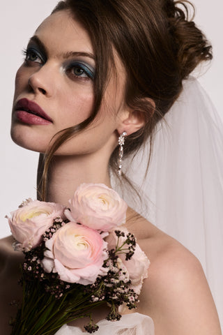 A MODEL LOOKING UPWARD WEARING THE MATERIAL PEARL LINEAR EARRINGS, A VEIL IN HER HAIR, AND HOLDING A BOUQUET OF FLOWERS.