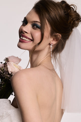 A MODEL LOOKING OVER HER SHOULDER WITH HER BACK TO THE CAMERA, WEARING THE MATERIAL PEARL LINEAR EARRINGS, A VEIL IN HER HAIR, AND HOLDING A BOUQUET OF FLOWERS. 