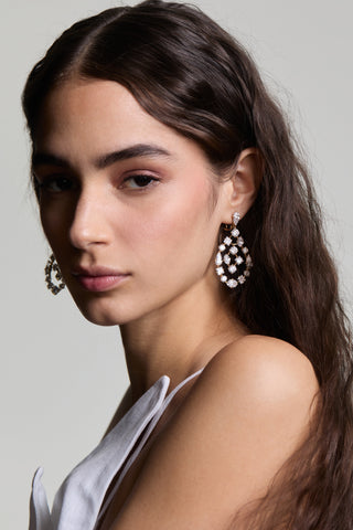 A MODEL LOOKING AT THE CAMERA WEARING THE RIVIERA CHANDELIER EARRINGS, WITH A STRAPPY TOP AND HER WAVY HAIR TUCKED BEHIND HER EAR