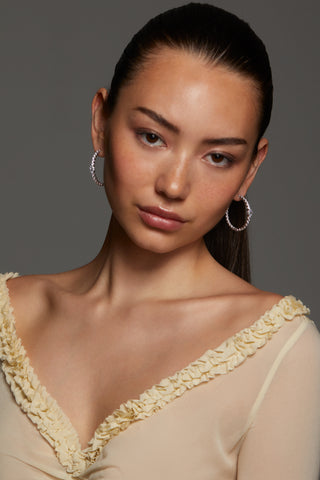 Model wearing a sheer beige top with floral details against a gray background. She is looking directly into the camera with her hair pulled back into a ponytail wearing STELLAR CZ HOOP EARRINGS