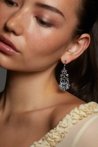 MODEL WEARING STELLAR MULTI STONE FEATHER DROP EARRINGS. IT IS A CLOSE UP IMAGE WITH THE MODEL GLANCING DOWN. SHE IS WEARING A YELLOW SHEER LONG SLEEVE DRESS WITH HER HAIR PULLED BACK INTO A PONYTAIL.