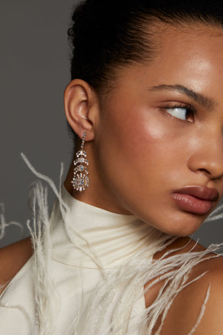 MODEL WEARING STELLAR FEATHER DROP EARRINGS. IT IS A CLOSE UP SHOT SHE IS LOOKING AWAY FROM THE CAMERA AND WEARING A TURTLENECK SLEEVELESS WHITE FEATHER TOP WITH HER HAIR PULLED BACK IN A BUN.