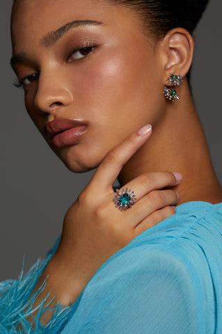 MODEL WEARING STELLAR MULTI STONE HALO RING. SHE HAS ON A BLUE DRESS WITH FEATHER SLEEVES AND HAIR PULLED UP INTO A SLEEK BUN. HER HAND IS REACHING TOWARDS HER FACE AS SHE LOOKS DIRECTLY INTO THE CAMERA. 
