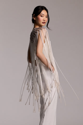 A MODEL WEARING WHITE TROUSERS AND A WHITE TOP WITH THE PEARL AND CZ CAPE DRAPED OVER HER SHOULDERS, TURNED TO THE SIDE AND LOOKING BACK OVER HER SHOULDER WITH HER HANDS IN HER POCKETS