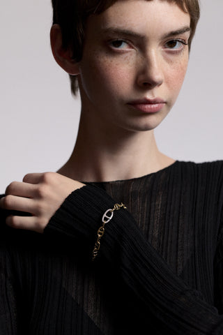 A MODEL LOOKING AT THE CAMERA WITH HER HAND RESTING ON THE OPPOSITE SHOULDER, AS SHE WEARS A SHEER BLACK DRESS AND THE CELINE ANCHOR LINK BRACELET