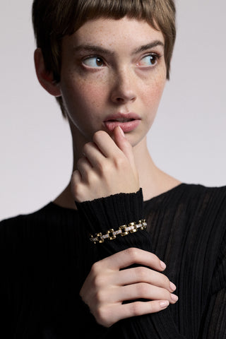 A MODEL LOOKING OFF TO THE SIDE WITH HER HAND LIFTED UP TO HER CHIN, AS SHE WERARS A SHEER BLACK TOP AND THE CELINE WATCH CHAIN BRACELET LAYERED OVER HER SLEEVE