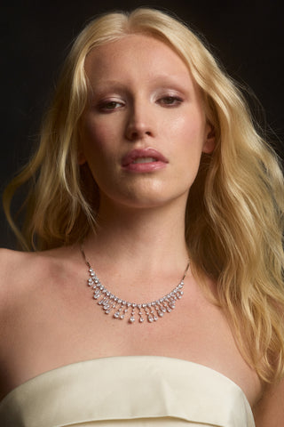 A MODEL WITH BLONDE HAIR LOOKING AT THE CAMERA, WEARING A WHITE STRAPLESS DRESS AND THE HERA FRINGE FRONTAL NECKLACE