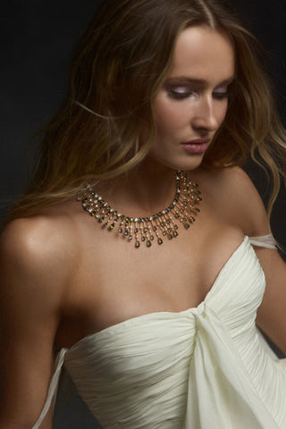 A MODEL LOOKING DOWN WITH HER BLONDE HAIR FLOWING AROUND HER, AS SHE WEARS A WHITE STRAPLESS DRESS AND THE HERA MULTI DRAMA COLLAR NECKLACE