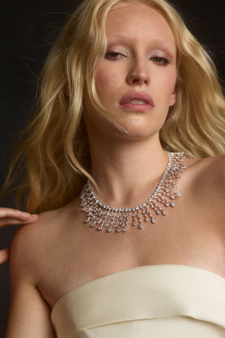 A MODEL LOOKING AT THE CAMERA WITH HER BLONDE HAIR BLOWING AROUND HER, WEARING A STRAPLESS WHITE TOP AND THE HERA DRAMA COLLAR NECKLACE