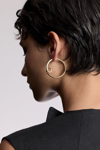 THE BLACK OF A MODEL’S HEAD TURNED SLIGHTLY TO THE SIDE, WITH SHORT BLACK HAIR WEARING THE MUSE SWIRL HOOP EARRINGS