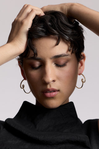 A MODEL WITH HER HANDS LIFTED UP TO HER HEAD AND HER EYES CLOSED, AS SHE WEARS A HIGH-NECK BLACK TOP AND THE MUSE SWIRL HOOP EARRINGS