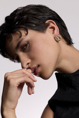 A MODEL TURNED SLIGHTLY TO THE SIDE LOOKING AT THE CAMERA WITH HER HAND LIFTED UP TO HER MOUTH, AS SHE WEARS A BLACK TOP AND THE MUSE SPHERE BUTTON EARRINGS
