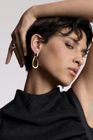 A MODEL TURNED SLIGHTLY TO THE SIDE WITH HER ARM RAISED AND DRAPED OVER HER HEAD, AS SHE WEARS A BLACK TOP AND THE MUSE DOOR KNOCK EAR EARRINGS