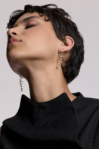 A MODEL WITH SHORT BLACK HAIR LOOKING UPWARDS WITH HER EYES CLOSED, AS SHE WEARS A BLACK TOP AND THE MUSE TWIRL LINEAR DROP EARRINGS