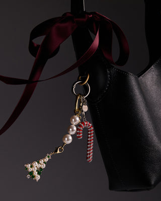 Candy Cane shaped bag charm and Christmas tree shaped bag charm with pearl, cubic zirconia, and green stone embellishments hanging from a pearl chain shown attached to a black handbag against a dark background.