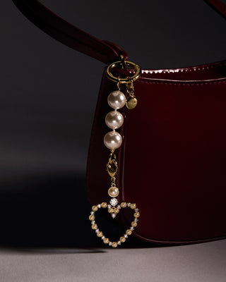 Pearl heart bag charm attached to a dark burgundy handbag on a gray surface with a black background
