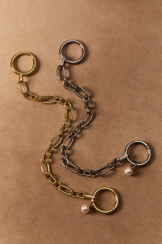 A SILVER-TONED BON VOYAGE CHAIN BAG SWAG 3 AND A GOLD-TONED BON VOYAGE CHAIN BAG SWAG 4, LAYING AGAINST A BROWN BACKDROP