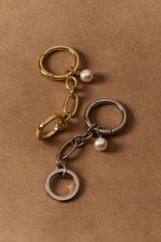 A SILVER-TONED BON VOYAGE PEARL CHAIN CHARM 3 AND A GOLD-TONED BON VOYAGE PEARL CHAIN CHARM 4 LAYING AGAINST A BROWN BACKDROP