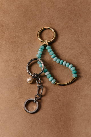 SILVER-TONED BON VOYAGE PEAL CHAIN CHARM 3 AND BON VOYAGE AMZ BAG CHARM 4 WITH GOLD-TONED HARDWARE, LAYING AGAINST A BROWN BACKDROP