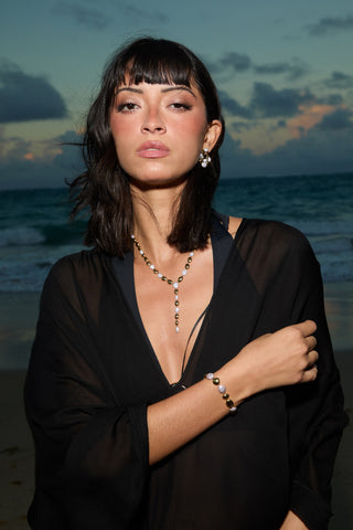 A MODEL ON A BEACH LOOKING AT THE CAMERA, WEARING A LOOSE BLACK COVER-UP, THE PEBBLES LINE BRACELET, AND THE PEBBLES Y NECKLACE