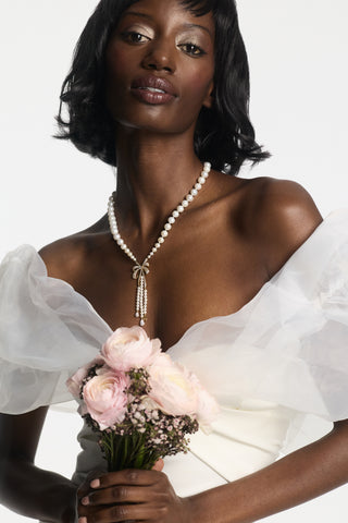 A MODEL WEARING THE EROS BOW PEARL NECKLACE AND WHITE GOWN, WHILE HOLDING A BOUQUET OF PINK FLOWERS