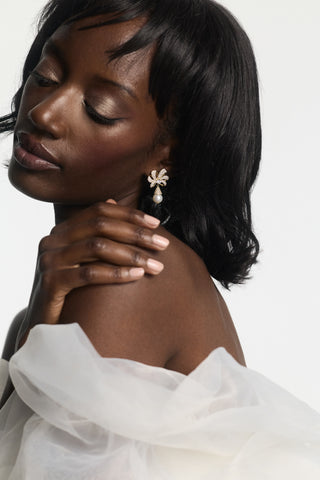 A MODEL LOOKING DOWN WITH HER HAND PLACED ON HER SHOULDER, WEARING THE EROS PEARL DROP POST EARRINGS AND A WHITE DRESS WITH CHIFFON SLEEVES