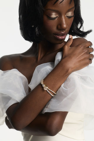 A MODEL IN A WHITE DRESS WITH CHIFFON SLEEVES LOOKING DOWN WITH HER ARMS WRAPPED AROUND HERSELF, WEARING THE EROS BOW TENNIS BRACELET