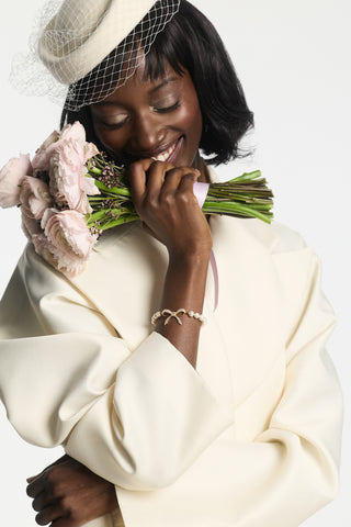MODEL WEARING THE EROS BOW AND PEARL LINE BRACELET WHILE HOLDING A BOUQUET, STYLED WITH A VEILED FASCINATOR AND CREAM BLAZER