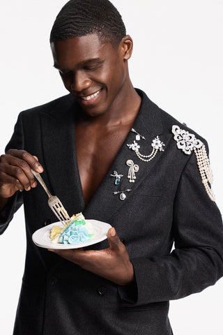 MODEL IN A SUIT JACKET EATING A PIECE OF CAKE, WITH CHARMS PINNED TO HIS LAPEL
