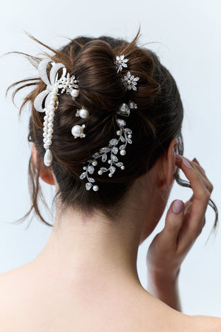 CLOSE-UP OF THE BACK OF A WOMAN’S HEAD WITH BRUNETTE HAIR STYLED IN A BUN, AS SHE BRUSHES HER BANGS AWAY FROM HER FACE, FEATURING MULTIPLE NADRI HAIR ACCESSORIES PINNED THROUGHOUT THE BUN.