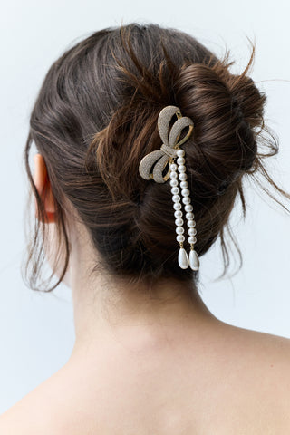 CLOSE-UP OF THE BACK OF A WOMAN’S HEAD WITH BRUNETTE HAIR STYLED IN A BUN, FEATURING NADRI’S BOW PEARL STRAND HAIR COMB PINNED INTO THE BUN WITH PEARL STRANDS CASCADING DOWNWARD.