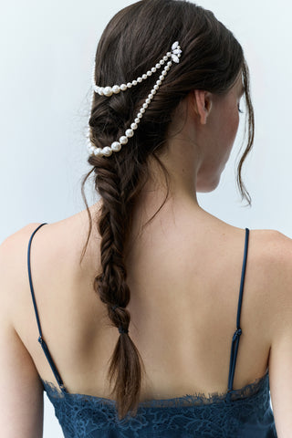 WOMAN IN A BLUE DRESS VIEWED FROM BEHIND, SLIGHTLY LOOKING DOWN WITH A LONG BRUNETTE BRAID BEHIND HER, FEATURING NADRI’S PEARL SWAG HAIR PIN PINNED ATOP HER HEAD