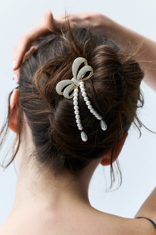 CLOSE-UP OF THE BACK OF A WOMAN’S HEAD WITH BRUNETTE HAIR STYLED IN A BUN, FEATURING NADRI’S BOW PEARL STRAND HAIR COMB PINNED INTO THE BUN WITH PEARL STRANDS CASCADING DOWNWARD.