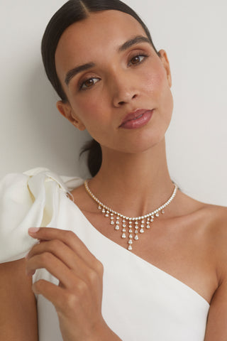 CLOSE UP IMAGE OF MODEL WEARING THE WILLOW CZ DRAMA DROPS COLLAR NECKLACE WITH A WHITE OFF THE SHOULDER BRIDAL DRESS WITH BOW DETAIL.  HER HAND IS TOUCHING HER SHOULDER AS SHE GAZES INTO THE CAMERA WITH A SLIGHT HEAD TILT AND A SOFT SMILE. 