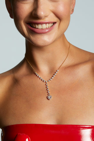 CLOSE UP IMAGE OF MODEL SMILING AND WEARING THE LE BISOU HALO HEART Y NECKLACE WITH HER HAIR UP IN A BUN AND WEARING A RED STRAPLESS DRESS.